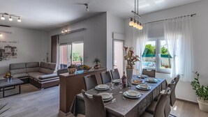 A modern dining area with a long table set for six, surrounded by chairs. A sofa is visible in the background, and there is a large window with sheer curtains. - Beautiful home in Marcana (Marcana)