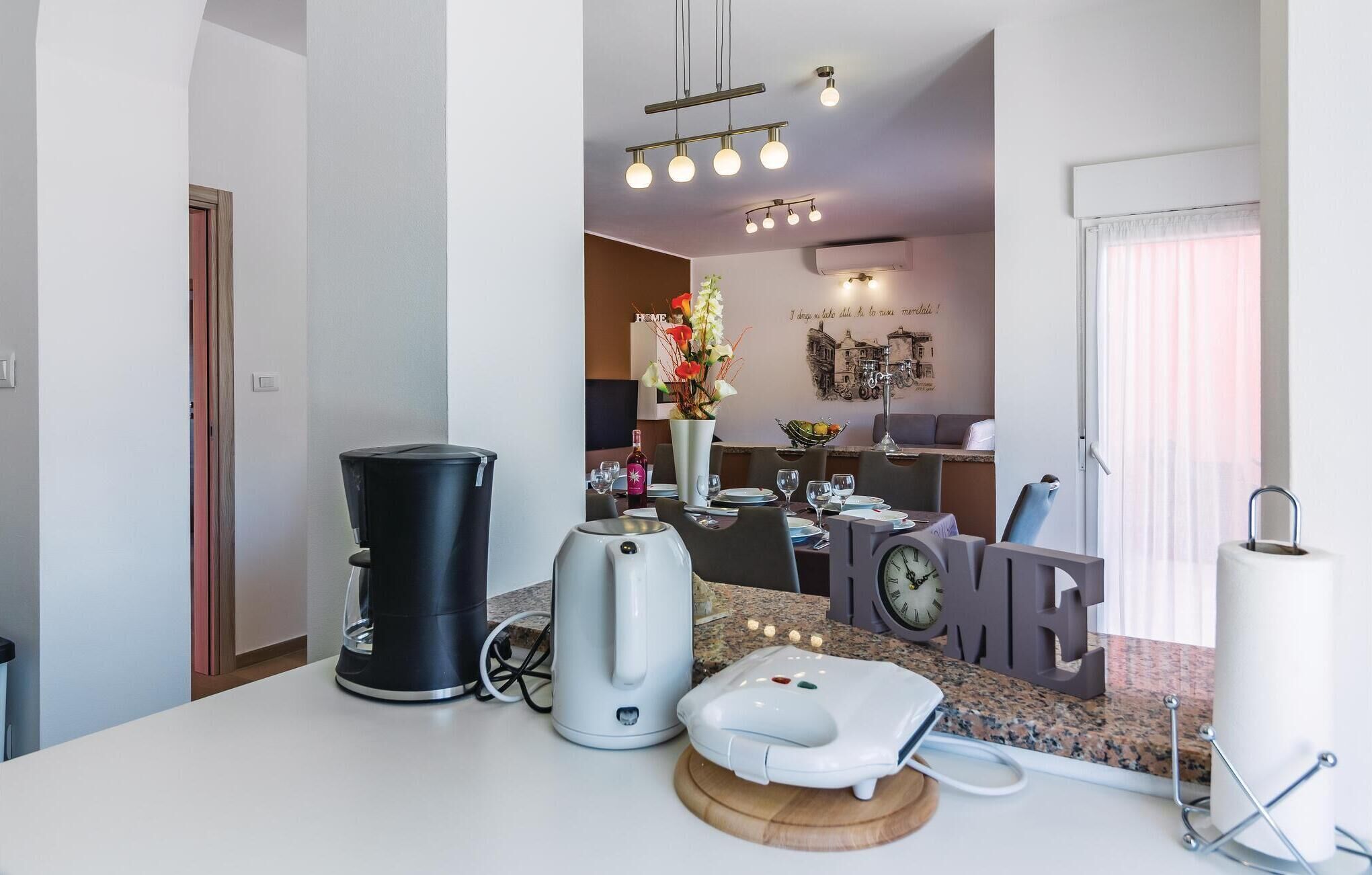 Une cuisine moderne équipée d’une machine à café, d’une bouilloire et d’un grille-pain sur le plan de travail. On y trouve également un coin repas avec une table dressée pour deux personnes, ainsi qu’un mur décoratif agrémenté d’une horloge et d’un panneau.