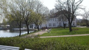 Un parc avec un bâtiment blanc, des bancs et un sentier qui longe un plan d’eau.
