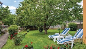 Um jardim com gramado, duas espreguiçadeiras listradas, uma mesa pequena com uma garrafa e um copo, e plantas em vasos.
