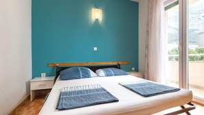 A bedroom with a teal wall, a wooden headboard, two bedside tables, a bed with a white mattress and two blue blankets, and a window with a view of greenery. - Lovely apartment in Makarska with WiFi (Makarska)
