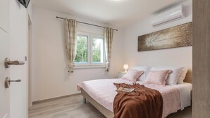 A bedroom with a bed, a window with curtains, an air conditioning unit, and a wall-mounted artwork.