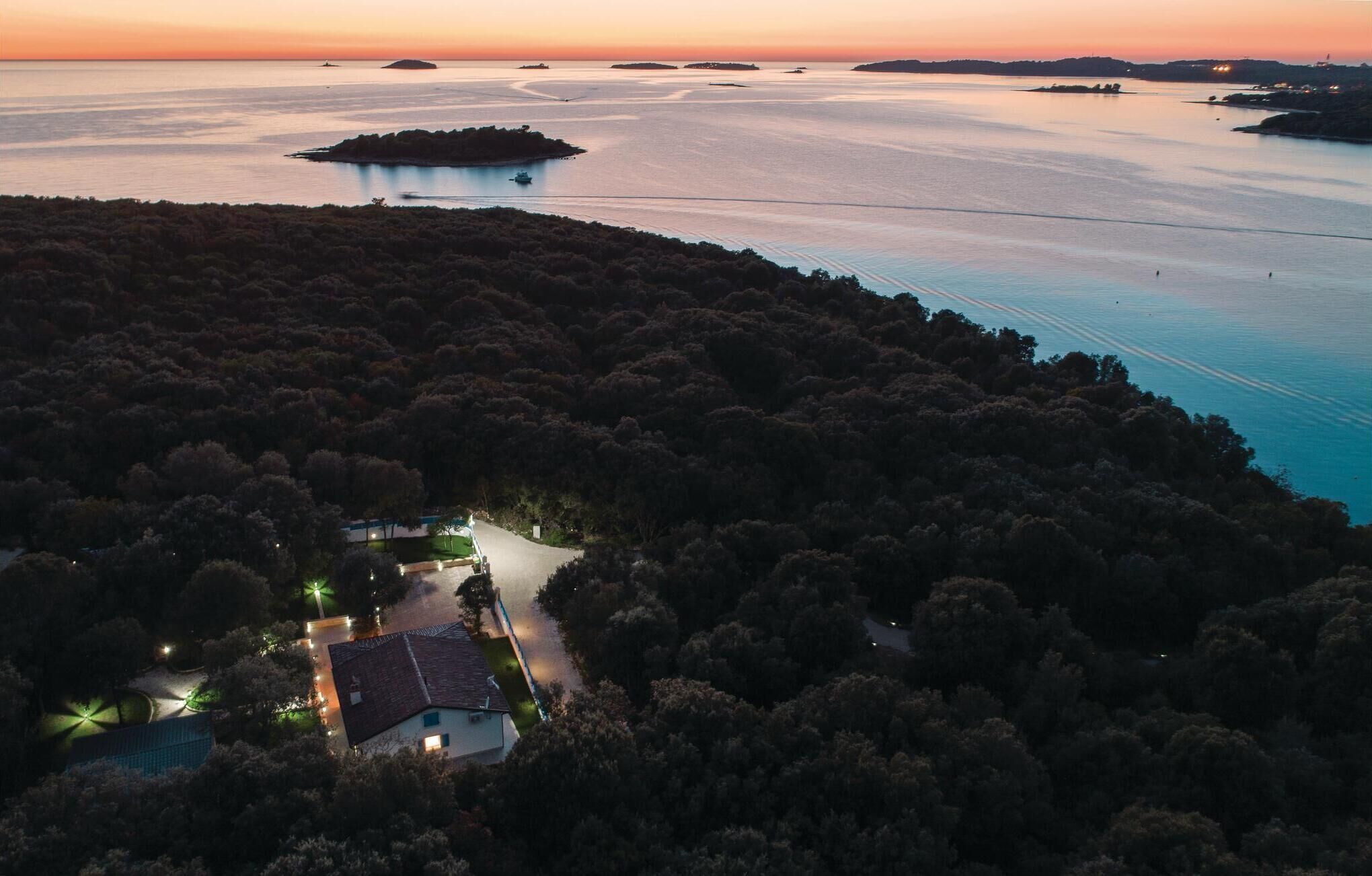 Une vue aérienne d’une zone côtière, avec une maison entourée d’une forêt dense, un ciel dégagé et des eaux calmes où naviguent des bateaux.