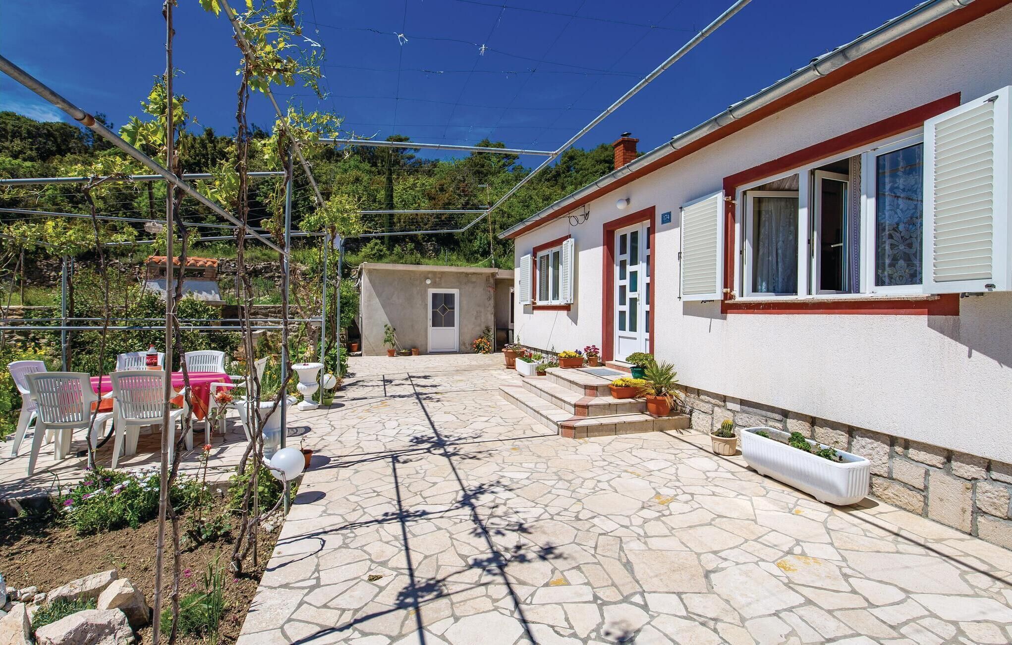 Une cour avec un sol en pierre, des chaises blanches, des plantes en pot et un bâtiment blanc orné de bordures rouges.