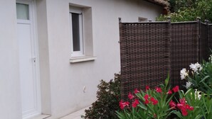 A white building with a tiled roof, a small window, and a door. There is a brown fence and some greenery with pink flowers. - Maisonette classified ***, in the heart of the greenery, between ocean and Landes forest (Saint-Julien-En-Born)