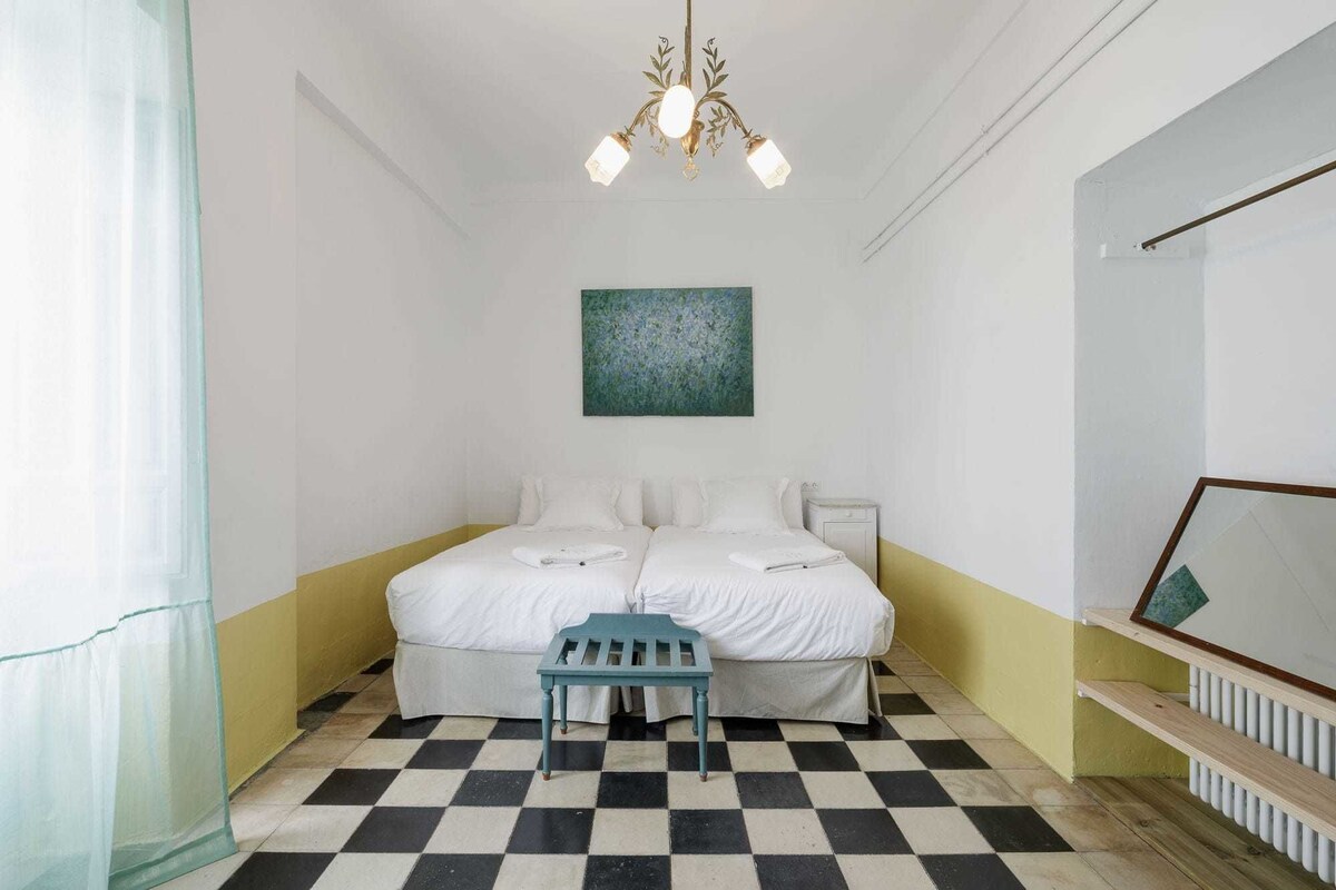 Habitación de hotel con piso a cuadros, cama con sábanas blancas, un pequeño banco azul, un cuadro en la pared y un candelabro.
