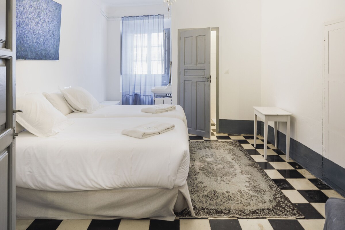 Habitación de hotel con dos camas, suelo a cuadros, araña y ventana con cortinas azules, además de una mesita.