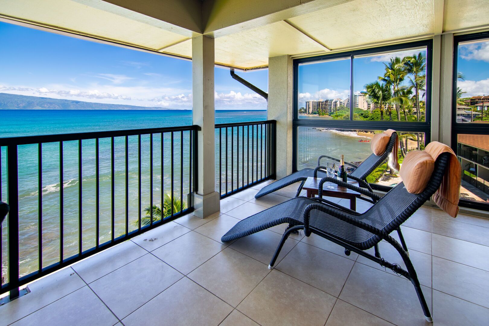 A balcony with two lounge chairs, a table, and a bottle of champagne. The balcony overlooks a beach with palm trees and buildings in the background.