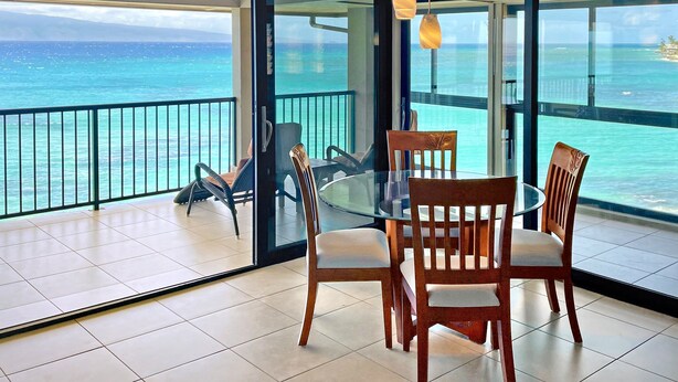 A balcony with a glass table and chairs, overlooking a turquoise sea and distant land.
