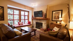 A cozy living room with leather sofas, a stone fireplace, a television mounted above it, and a large window with a view of snow-covered trees. - Spacious Rustic Whistler Retreat at the Woods (Whistler)