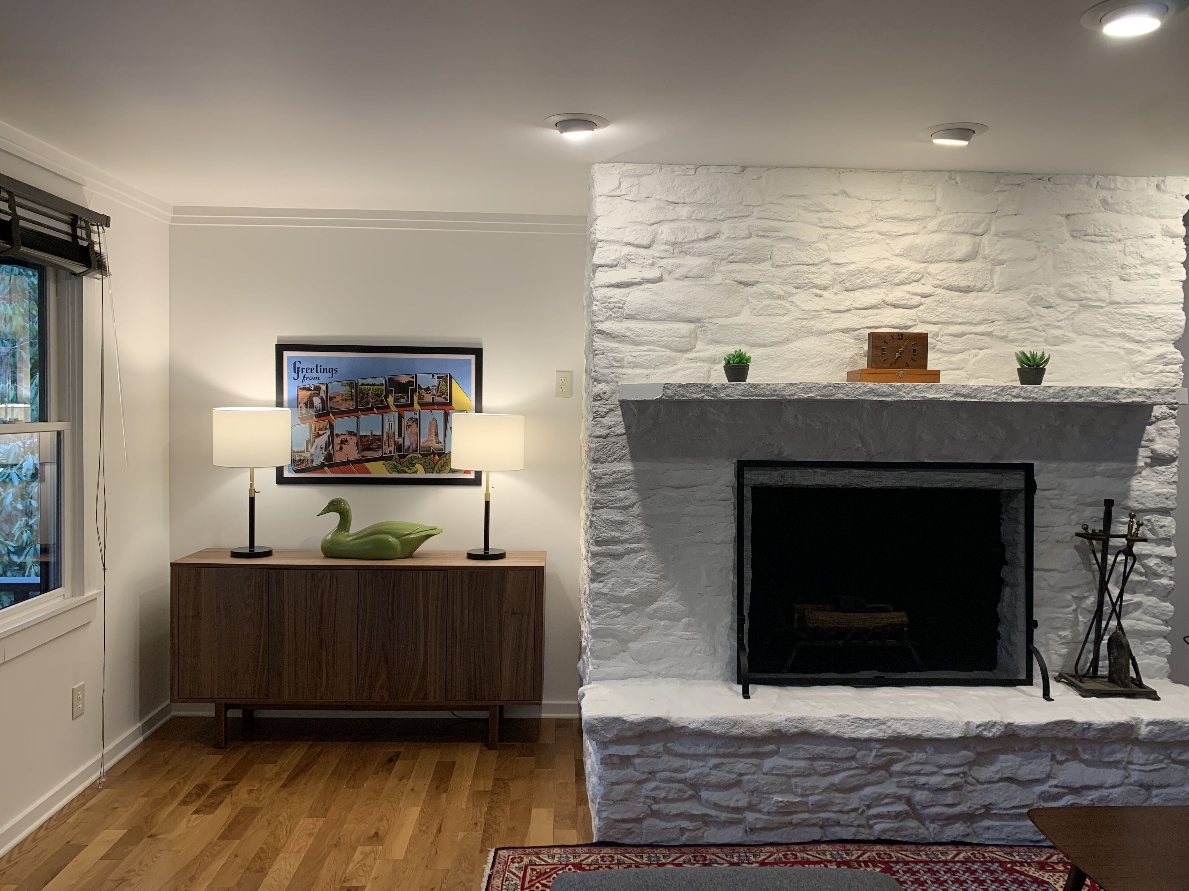 A living room with a white stone fireplace, a wooden console table with a green swan decoration, and a framed picture of postcards.