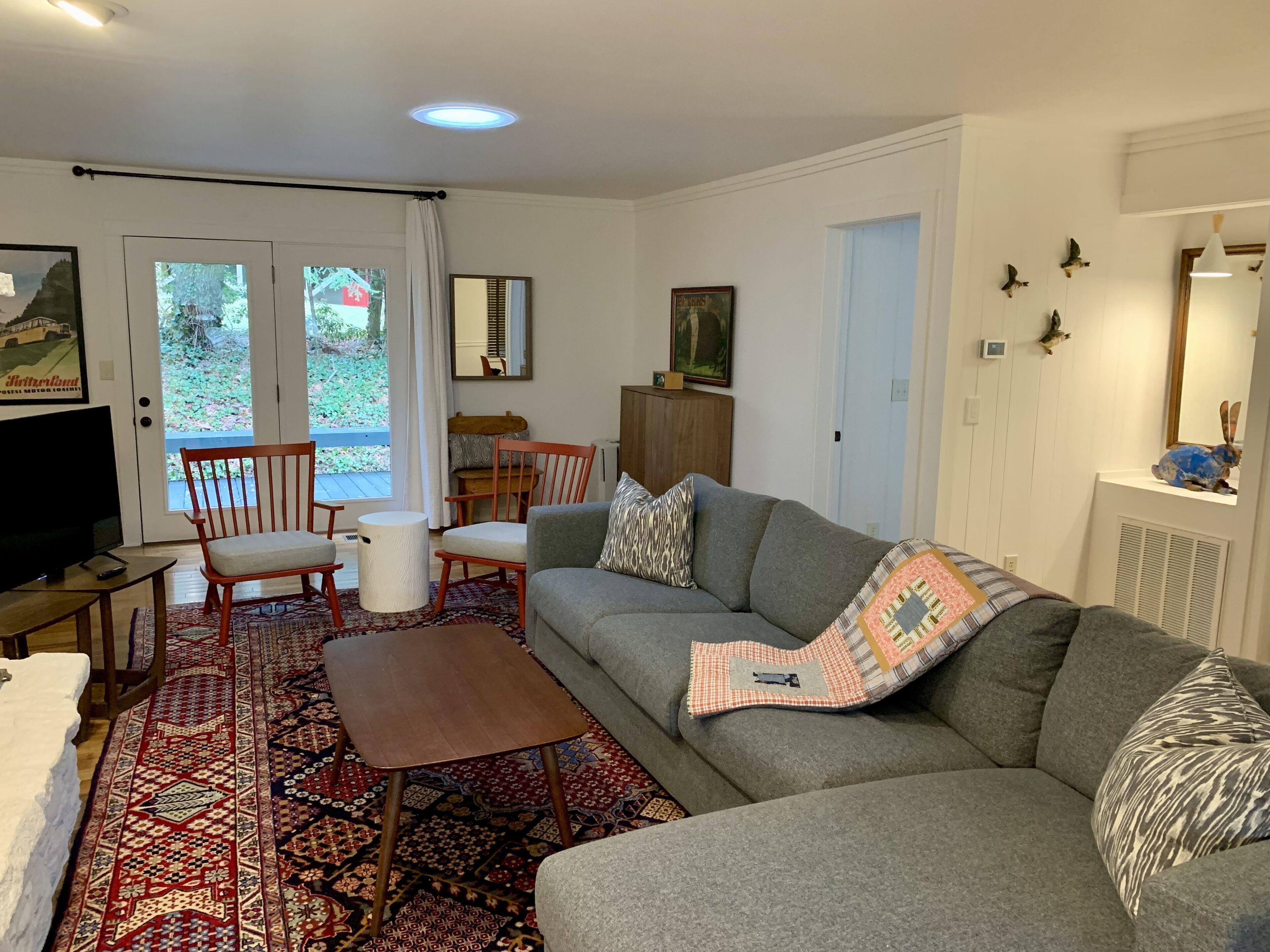 A living room with a grey sofa, a wooden coffee table, and a patterned rug. There is a television on a wooden stand, a chair, and a cabinet.