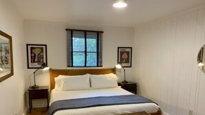 Un dormitorio ordenado con una cama de madera, dos mesitas de noche con lámparas, una ventana con persianas y una alfombra estampada.