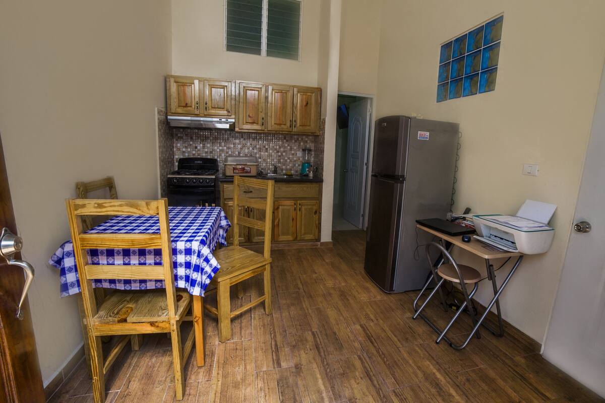 A compact kitchen with wooden cabinets, a gas stove, a refrigerator, and a table with a checkered tablecloth.