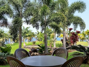 An outdoor seating area with a round table and chairs, surrounded by various tropical plants and palm trees, with a view of the sea in the background. - Oceanfront Vacation Rental (Hakalau)