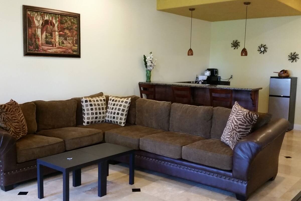 A living room with a brown sectional sofa, a black coffee table, and a painting of a garden scene on the wall.