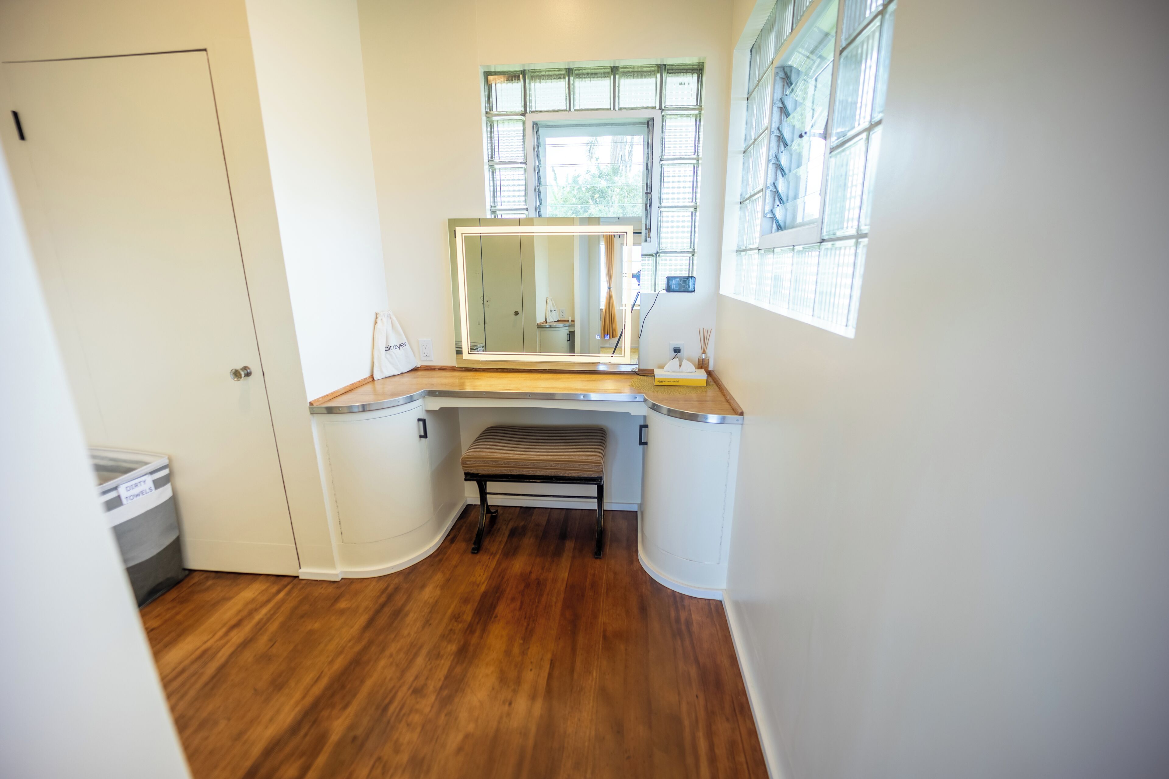 Une petite salle de bain avec un plancher en bois, un banc, un grand miroir et une fenêtre avec des stores.