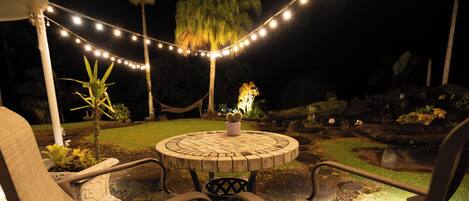 An outdoor seating area with a round table and chairs, surrounded by string lights and plants.