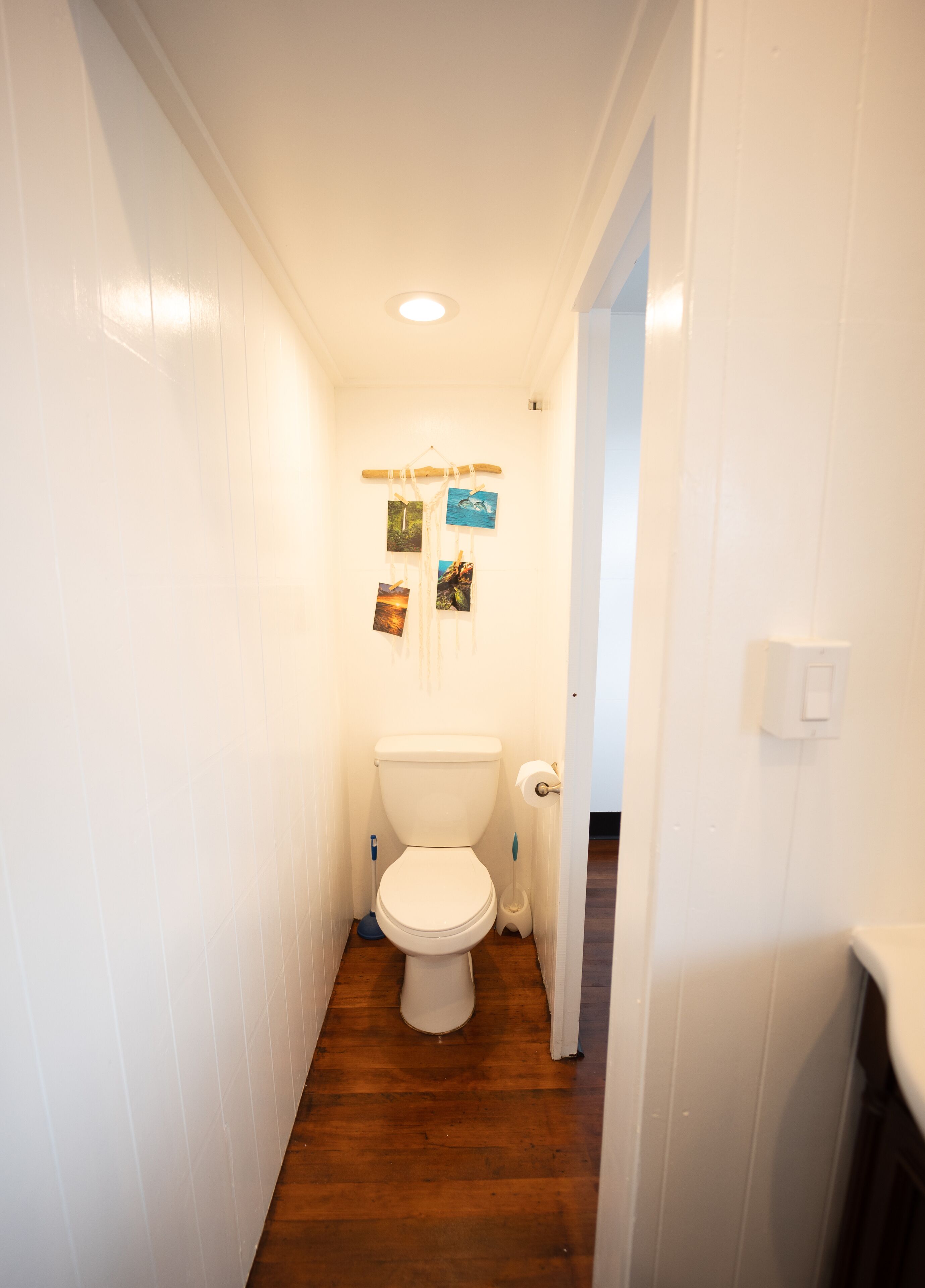 Une petite salle de bain aux murs blancs, avec un plancher en bois et des toilettes. Trois tableaux sont accrochés au mur, au-dessus des toilettes.