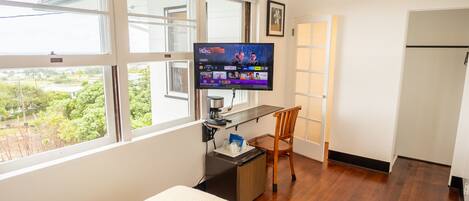 A hotel room with a bed, a desk with a computer, a chair, a television, and a window with a view of greenery.
