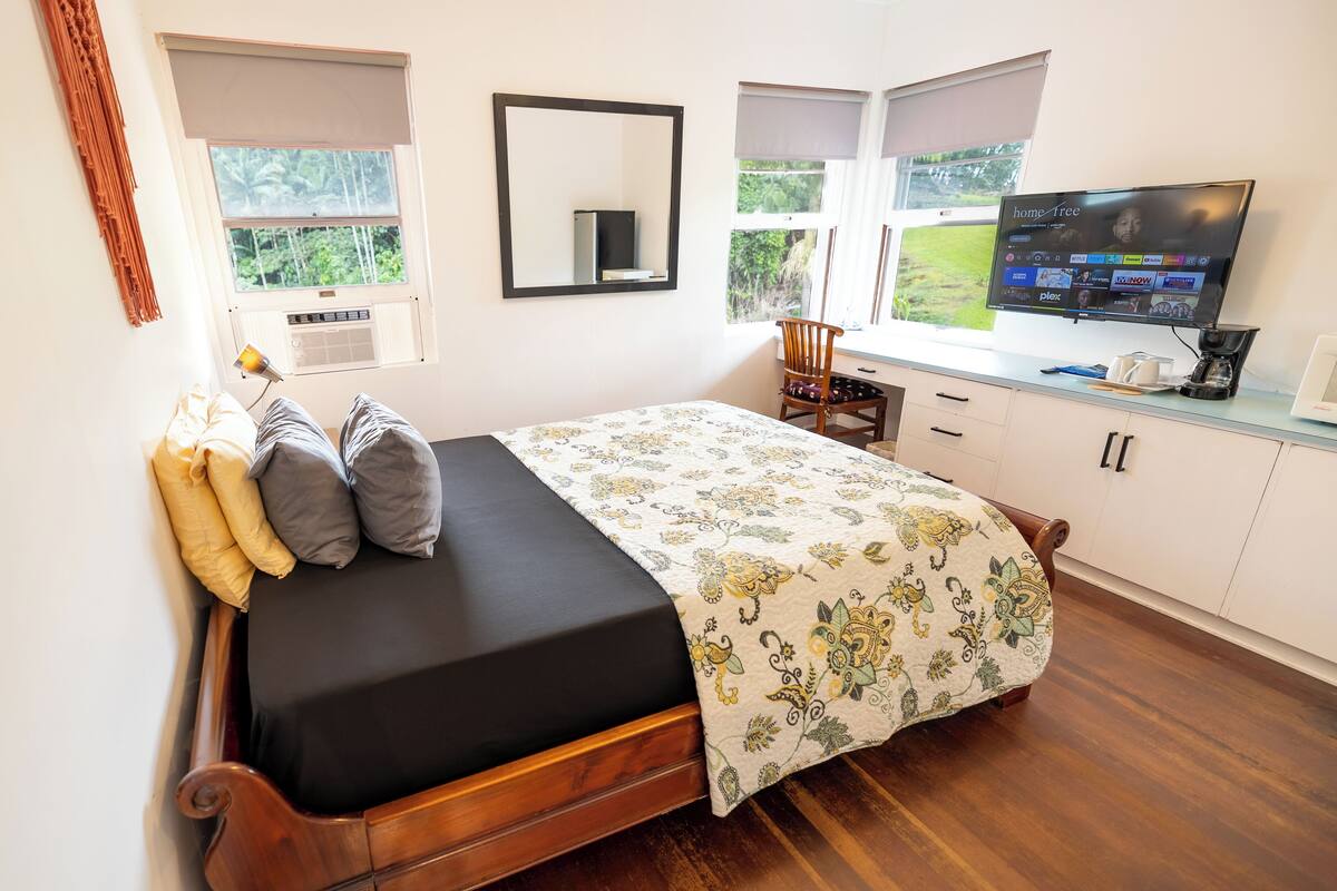 A bedroom with a bed, a desk, a chair, a television, and a window with a view of greenery.