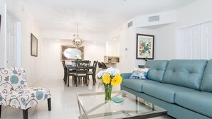 A living room with a blue sofa, a glass coffee table with a vase of flowers, a dining area with a chandelier, and a ceiling fan. - 1900 ft from beach Luxury Jupiter Beach Villa! private entrance community pool (Jupiter)