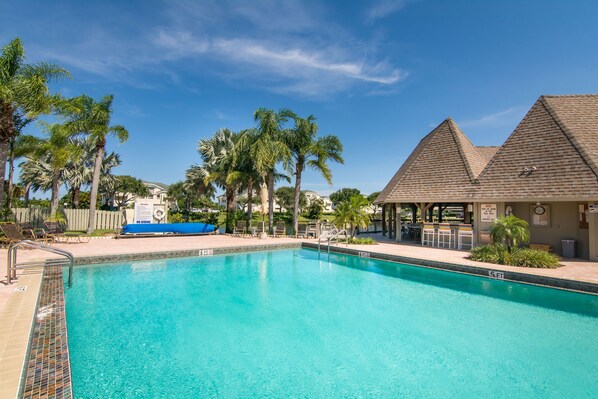 Una piscina azul cristalina con borde de azulejos, rodeada de palmeras y una zona cubierta con asientos.