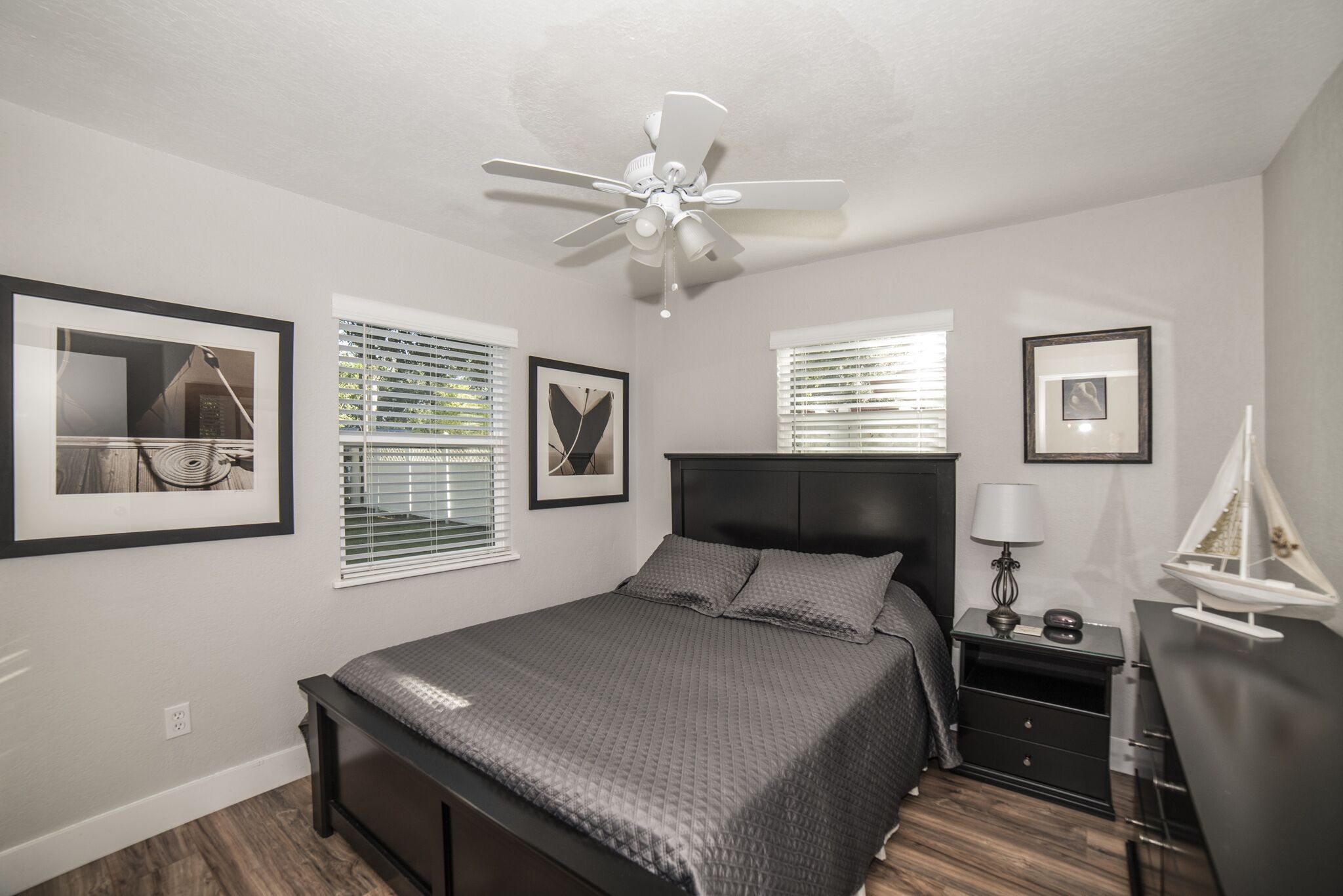 A bedroom with a bed, a nightstand, a lamp, a ceiling fan, and framed pictures on the walls.