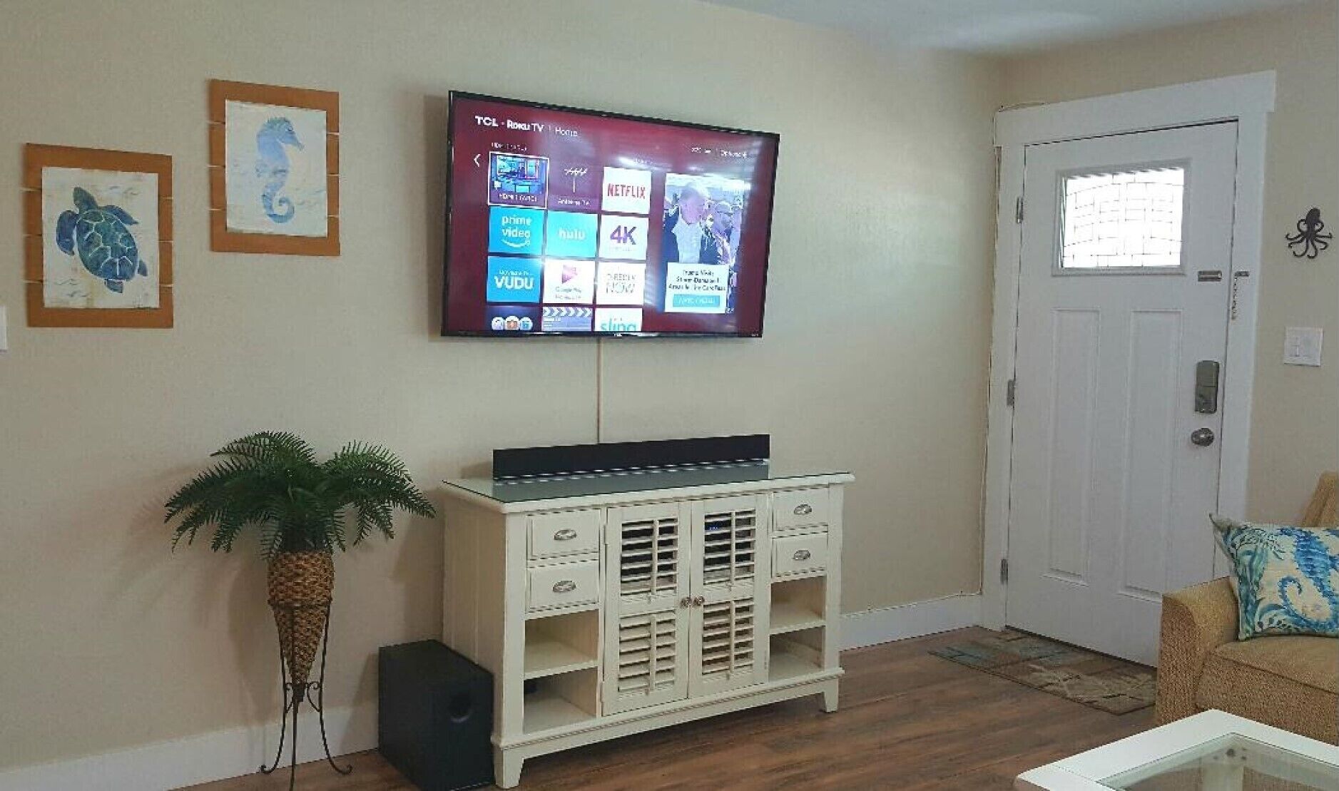 Un salón con un televisor de pantalla plana montado en la pared que muestra varios servicios de streaming. Hay un mueble blanco con cajones y una planta en maceta a su lado.