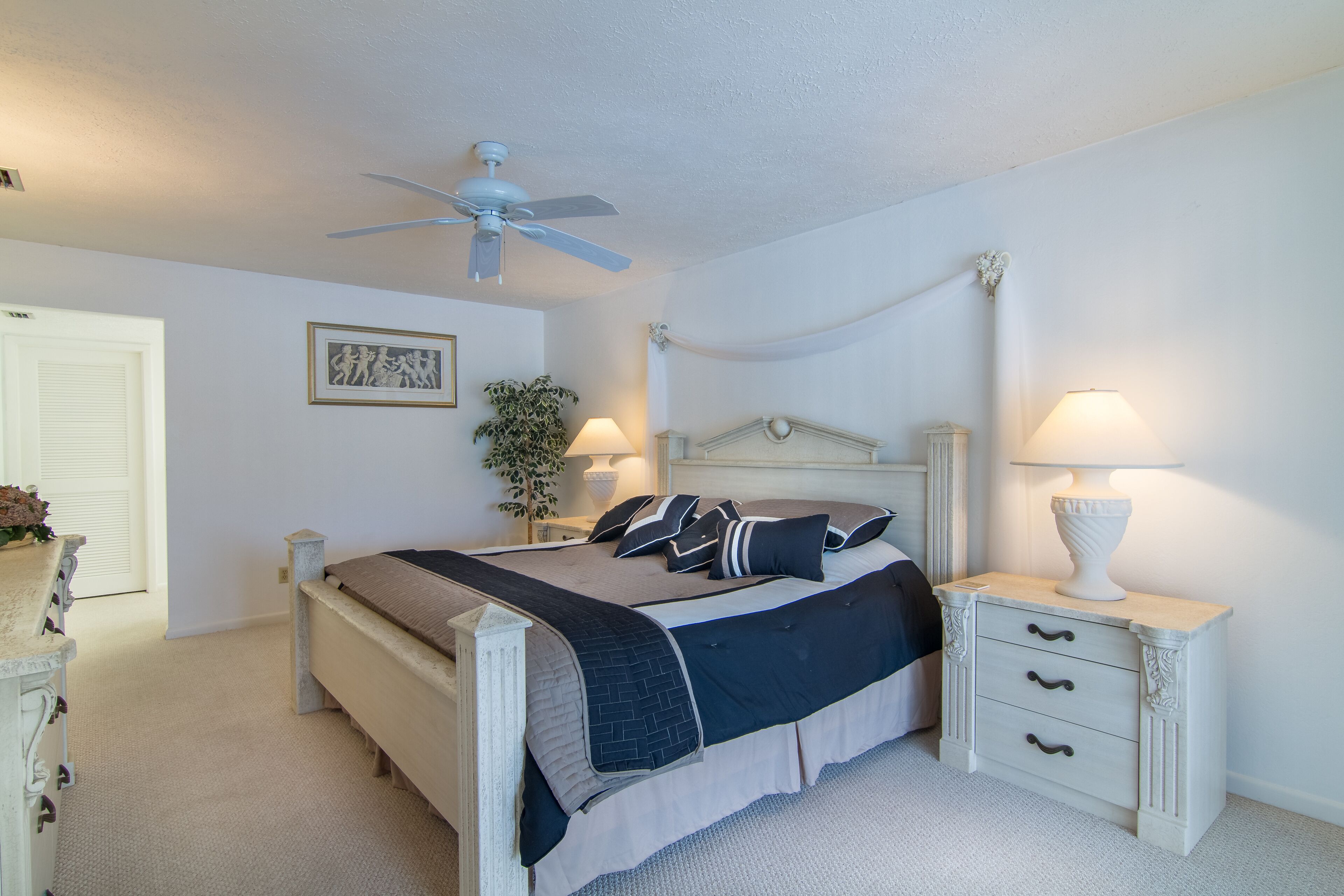 Une chambre à coucher comprenant un lit, des tables de chevet, un ventilateur de plafond et une petite table de nuit avec une lampe.