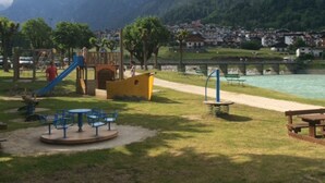 Ein Spielplatz mit Rutschen und Schaukeln, einem Picknicktisch und Blick auf einen Fluss und Berge im Hintergrund.