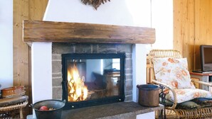 Une cheminée avec un foyer en pierre, une cheminée en bois et un fauteuil en osier agrémenté d’un coussin à motifs floraux placé à proximité.
