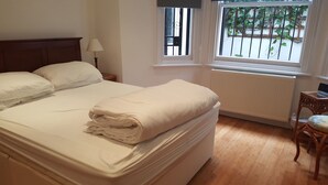 A neatly made bed with white linens, a wooden headboard, a bedside table with a lamp, a window with a view of greenery, and a radiator. - Bright Garden Flat w/ Patio | 5★ Reviews | 5 min to Tube & Westfield (London)