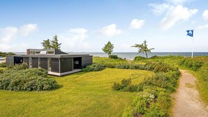 A modern house with a large deck, surrounded by lush greenery and a view of the sea.