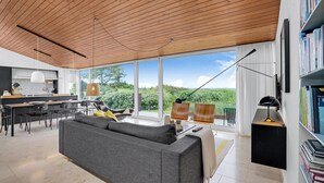 A modern living room with a large grey sofa, a dining area with a table and chairs, and a kitchen in the background.