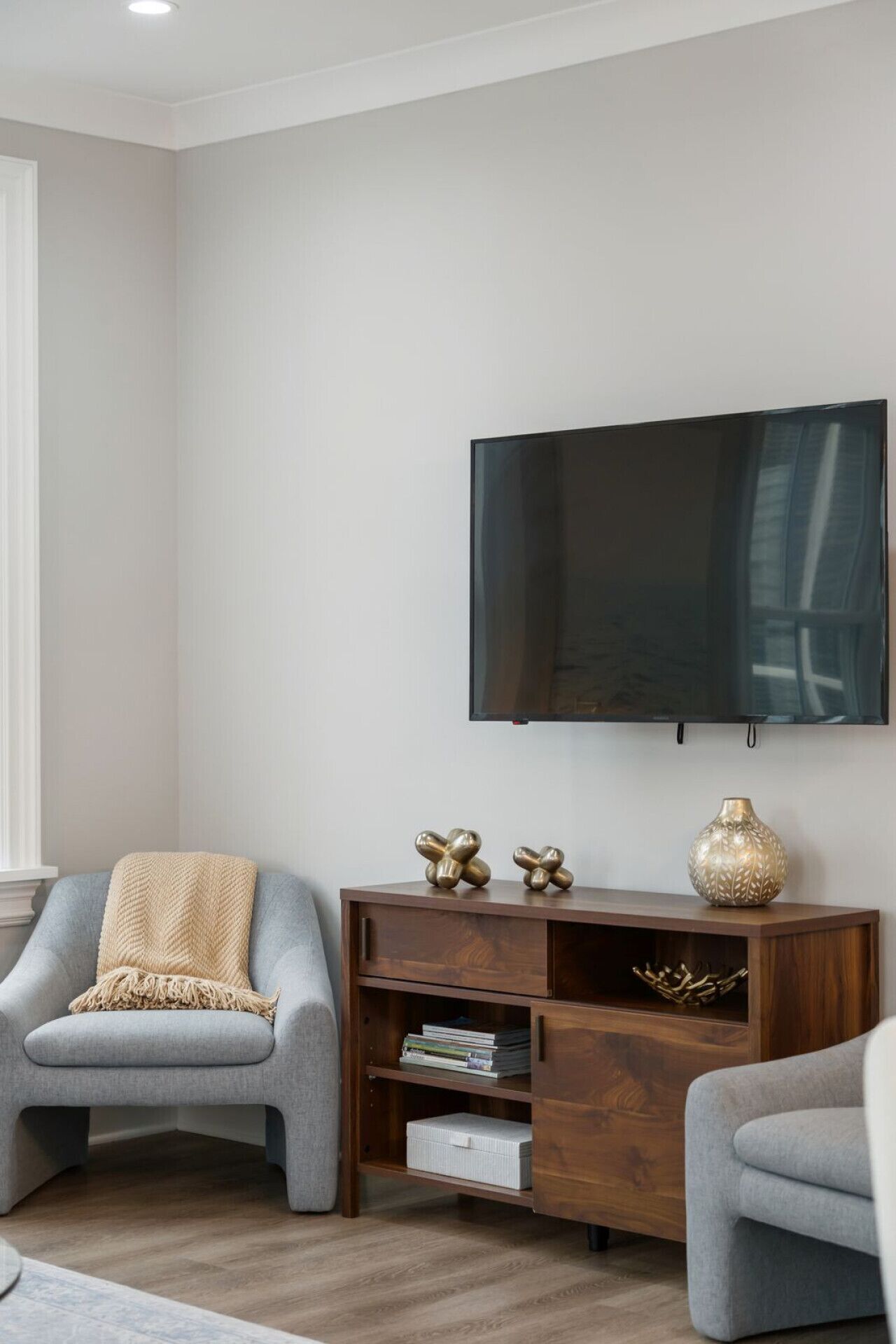 Una sala de estar con un televisor de pantalla plana montado en la pared, dos sillones grises, una mesita de madera con objetos decorativos y una estantería.