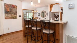 Una cocina con barra, taburetes de madera, luces colgantes, un reloj y un gallo decorativo.