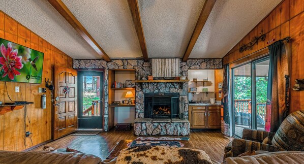 A cozy cabin interior with a stone fireplace, wooden walls, a dining area, and a large window. - Hidden Treasure | Strike Gold w/ Hot Tub Nights, Cozy Cabin Charm & Pet Friendly (Ruidoso)