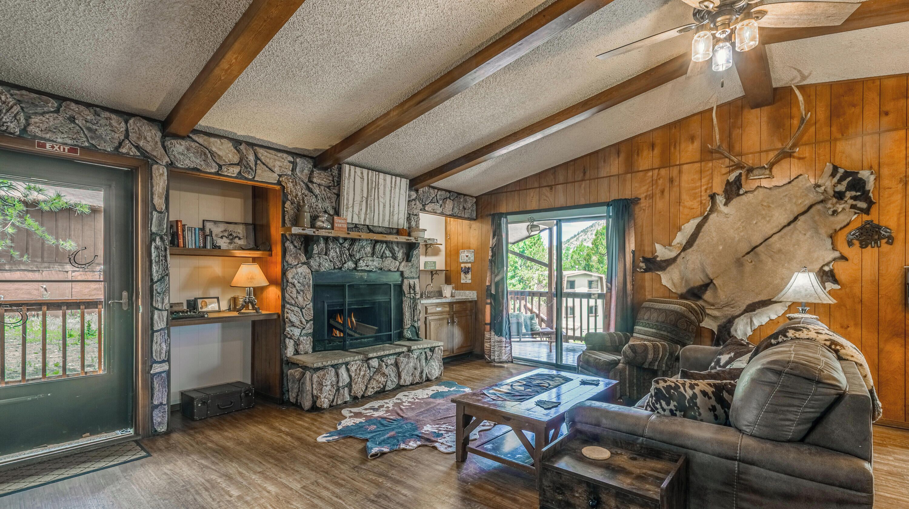 Un intérieur rustique avec une cheminée en pierre, des poutres en bois, un canapé en cuir et une peau d’animal fixée au mur.