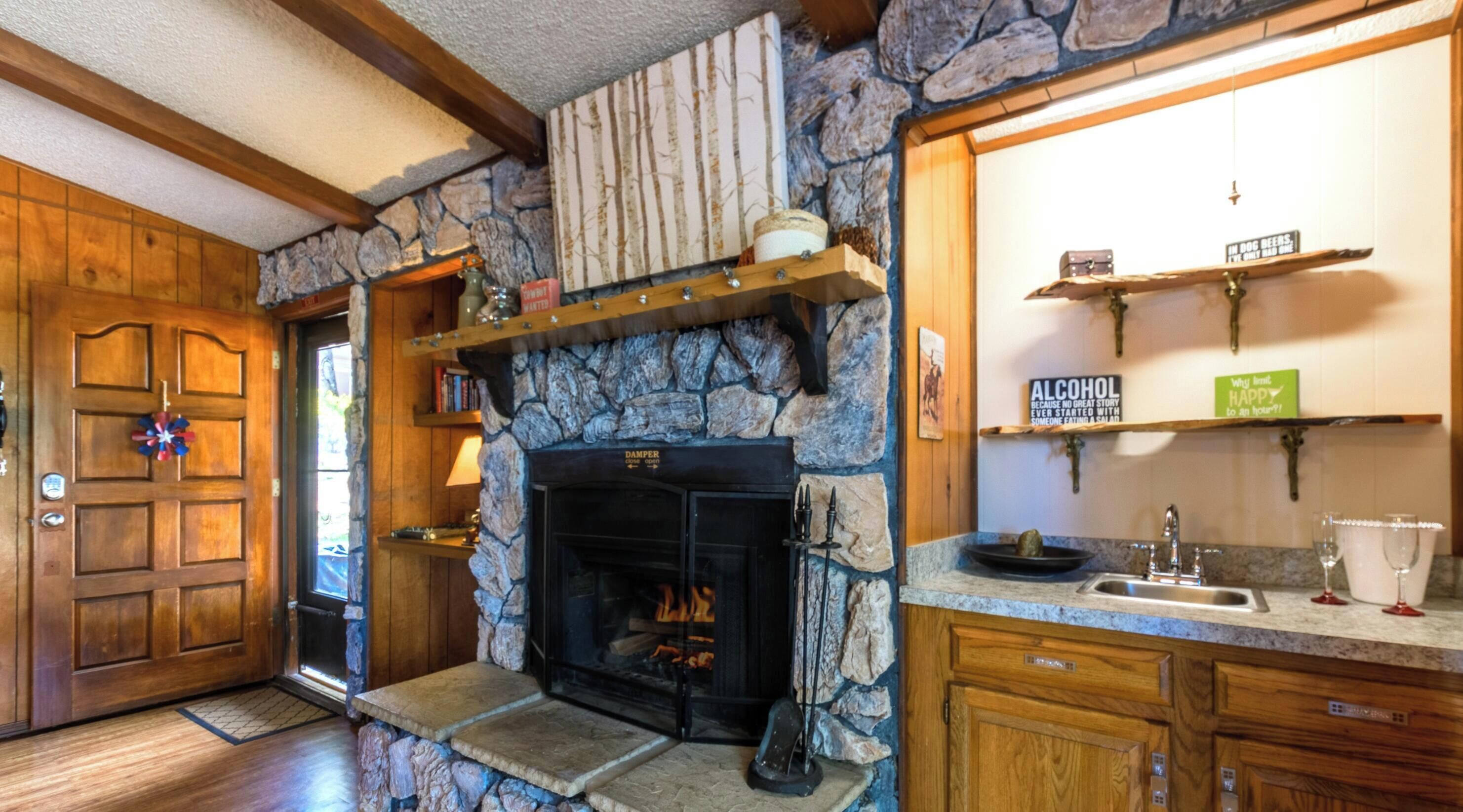 Une cheminée en pierre avec un registre, des étagères en bois au-dessus, une porte en bois ornée d’une guirlande décorative et un coin cuisine avec un évier et un plan de travail.
