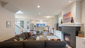 A modern living room with a dark-colored sofa, a fireplace, and a kitchen in the background. - Hillside Haven - Bend's West Side, Work-from-Home Office, New, Modern, Light, Bright (Bend)