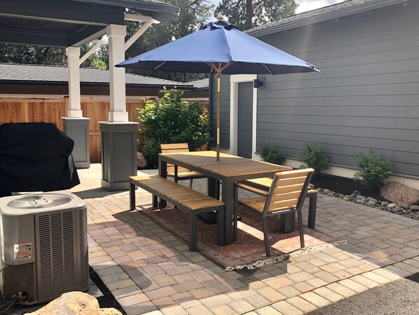 An outdoor patio with a wooden table and chairs, a blue umbrella, and a grill.