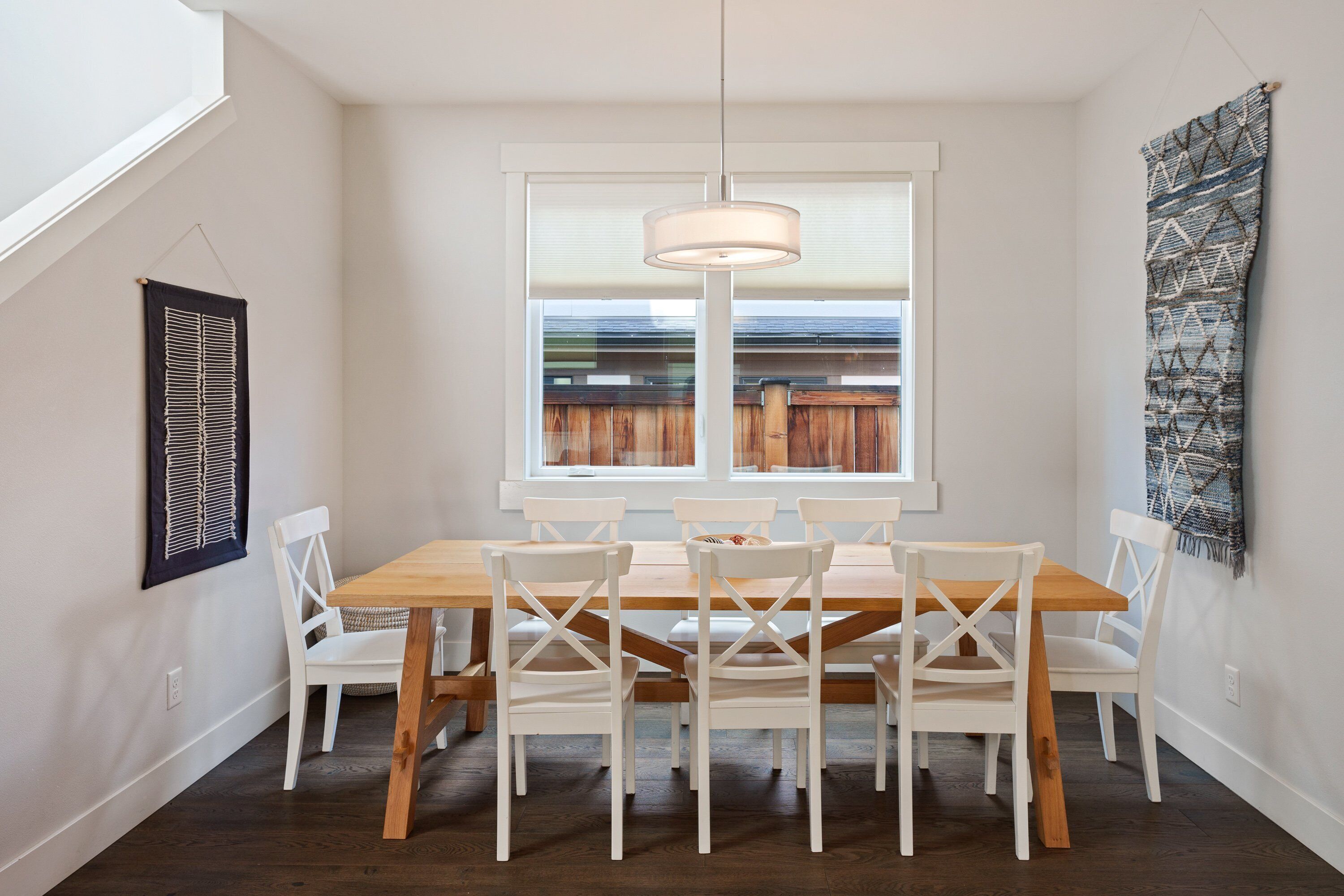 A dining area with a wooden table and white chairs, a hanging light fixture, and a window with blinds.