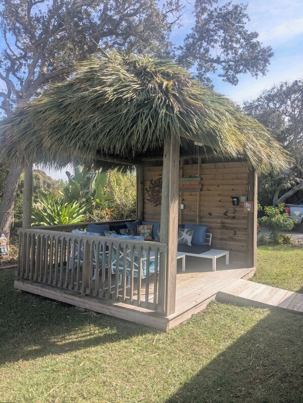 A wooden gazebo with a thatched roof, furnished with a sofa and a small table, surrounded by a wooden fence.