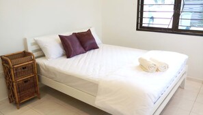 Una cama bien hecha con ropa de cama blanca y dos almohadas, una toalla enrollada al lado y una cesta de mimbre cerca.