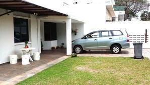Un coche aparcado en una entrada con un edificio blanco, una pequeña mesa y plantas en macetas.