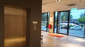 An elevator in a modern building with a digital display, a fire alarm, and a glass door leading to an outdoor area with a view of parked cars and a bus. - My Rainforest Suite Hotel (Langkawi)