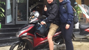 Dos personas de pie junto a una motocicleta roja estacionada frente a un edificio con amplias ventanas y una escalera visible en su interior.