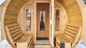 A wooden cabin with a central door, two benches on either side, and a fireplace visible through the door.