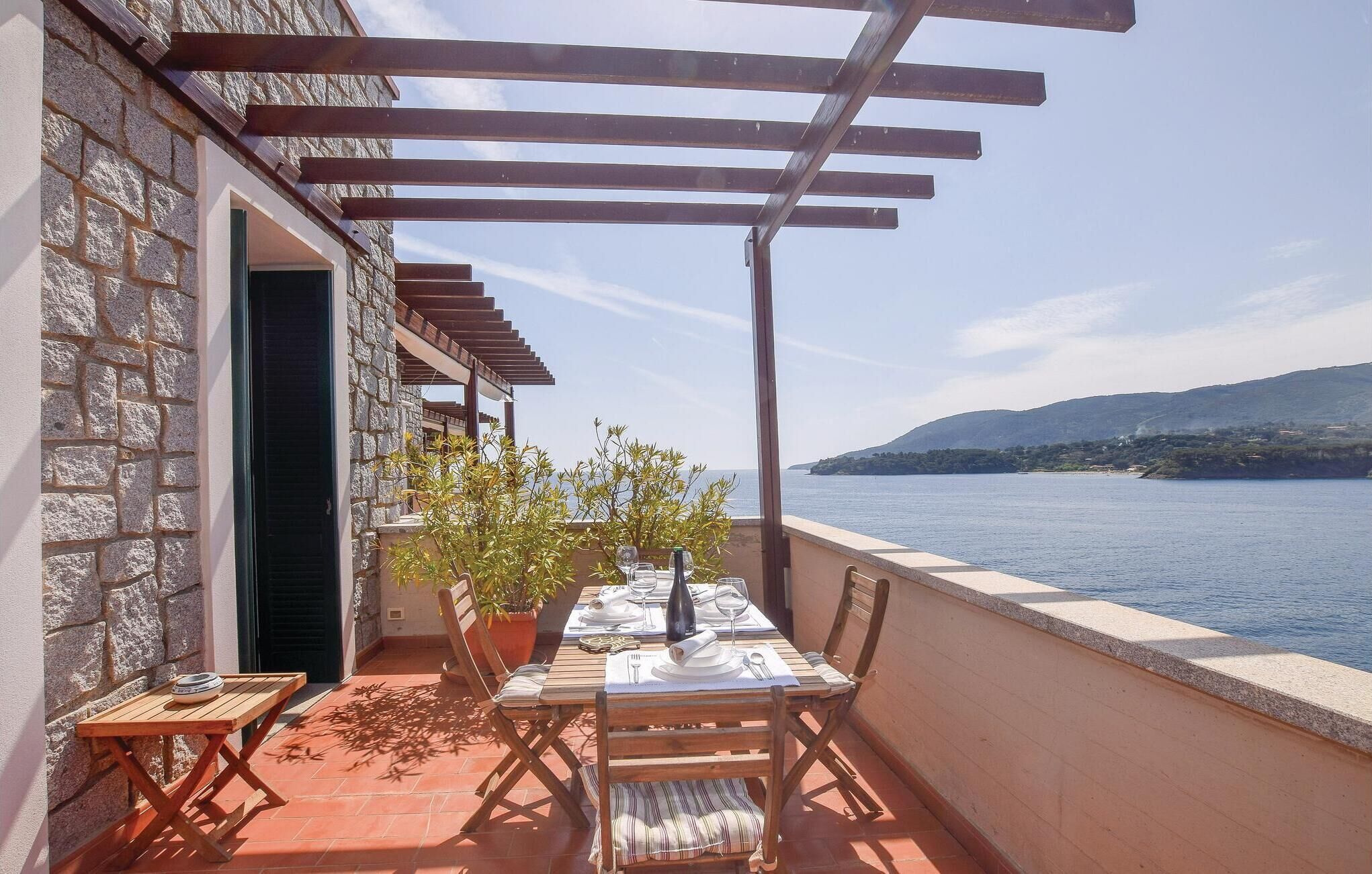 Un área de comedor al aire libre con una mesa lista para dos personas, con vistas a una masa de agua y una costa distante.